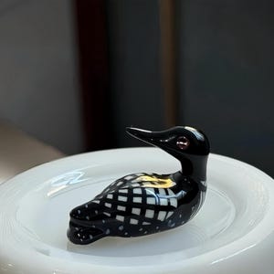 Könnte beinhalten: Eine dekorative Glasfigur eines Eistauchers. Der Vogel hat einen glänzend schwarzen Kopf und Hals mit einem roten Auge. Der Körper ist schwarz-weiß kariert. Die Figur ruht auf einer weißen, runden Oberfläche.