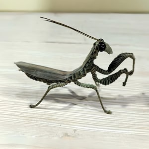 May include: A detailed metal sculpture of a praying mantis. The insect is crafted from thin, textured metal strips, with a body in shades of green and brown. It has long, slender legs and antennae, and large, dark eyes. The sculpture is posed on a light-colored wooden surface.