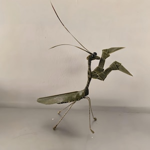 May include: A handcrafted praying mantis sculpture made from green and brown materials. The insect has detailed wings, a long neck, and wire legs. The sculpture is posed with its arms raised, showcasing its intricate design.