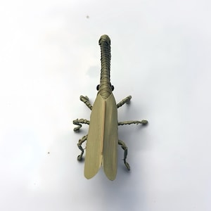 May include: A handcrafted insect figurine made from woven light green plant fibers. The sculpture features a long, slender neck, two prominent black bead eyes, and elongated, flat wing-like structures covering the back, with six thin, jointed legs.