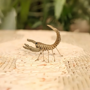 May include: A detailed, handcrafted scorpion sculpture in shades of tan and brown. The scorpion features intricate detailing on its segmented body, claws, and curved tail. The sculpture is posed on a textured surface, with a blurred green background.