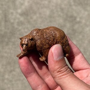 Könnte beinhalten: Eine kleine, braune Bärenfigur mit strukturierter Oberfläche in laufender Haltung. Der Bär weist detaillierte Fellzeichnungen, kleine abgerundete Ohren und dunkle Augen auf. Die Miniaturskulptur ist realistisch gestaltet und zeigt das Tier in einer nach vorne geneigten Körperhaltung, wobei die Vorder- und Hinterbeine so positioniert sind, als würde es sich über eine Oberfläche bewegen.