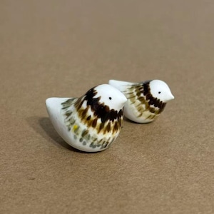 May include: Two small ceramic bird figurines featuring a white base color with brown and black feathered patterns. Each bird has a pointed beak, a small black eye, and a rounded body shape, showcasing a hand-painted aesthetic with visible brushstrokes.