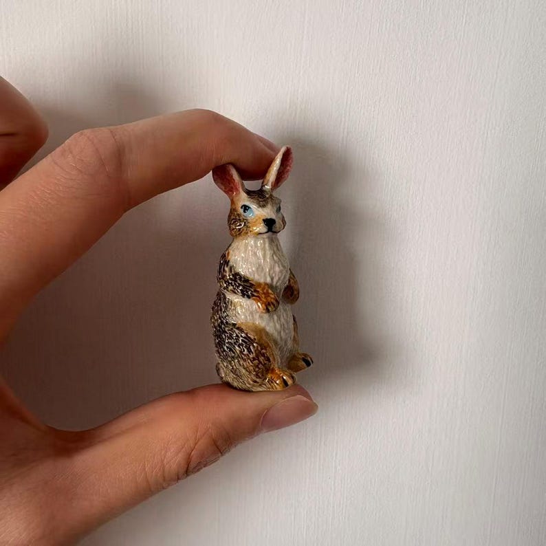May include: A small, detailed figurine of a rabbit. The rabbit has brown and white fur with blue eyes and long ears. The figurine is held between fingers against a white background.