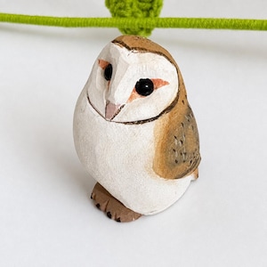 May include: A small hand-carved wooden barn owl figurine featuring a white face with black eyes, a light brown crown, and tan wings with subtle dark brown speckling. The owl has a rounded body shape and visible carved textures on its surface.