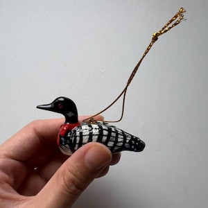 May include: A small loon ornament featuring a black head with a red eye, a red neck band, and a body patterned with black and white checkered markings. The bird figure includes a thin gold metallic string attached to the top for hanging purposes.