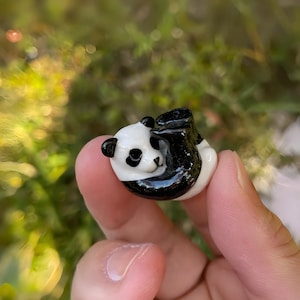 Może przedstawiać: Mała, błyszcząca figurka pandy w zabawnej pozie. Panda jest głównie biała z czarnymi oznaczeniami wokół oczu, uszu i ciała. Figurka trzymana jest w dłoni na rozmytym zielonym tle.