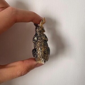 May include: A small, detailed figurine of a rabbit, held between fingers. The rabbit has a textured, speckled pattern in shades of brown, black, and tan. The ears are upright, and the overall design is realistic.