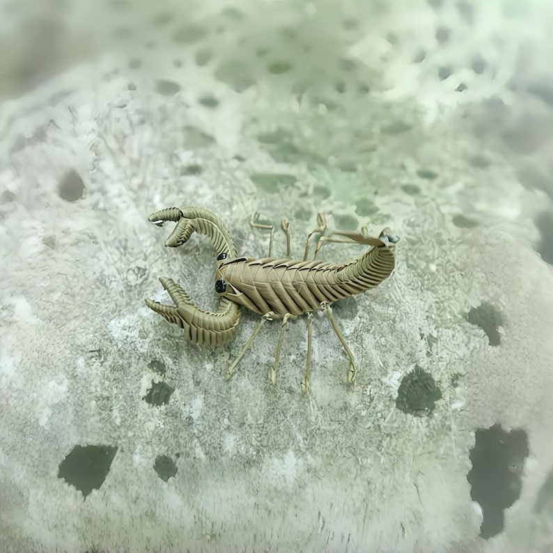 May include: A detailed, tan-colored scorpion figurine with articulated claws and segmented body. The scorpion is posed on a textured, light gray surface, creating a naturalistic scene. The figurine's design emphasizes its realistic features.