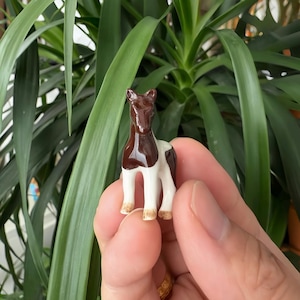 May include: A small ceramic horse ornament. The horse is brown and white, with a detailed design. The figurine is held in front of a backdrop of green foliage.
