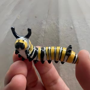 Könnte beinhalten: Eine Glas-Raupenfigur mit einem gelb-schwarz gestreiften Körper. Die Raupe hat ein weißes Gesicht mit schwarzen Antennen und schwarzen Füßen. Die Figur wird zwischen zwei Fingern gehalten.