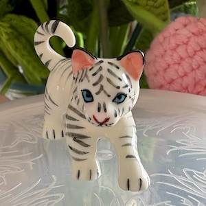 May include: A white ceramic tiger cub figurine with black stripes, blue eyes, and pink inner ears. The tiger is in a walking pose, with its tail curled. The figurine is on a light blue surface with a floral design.
