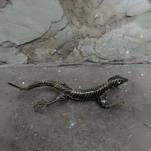 Könnte beinhalten: Eine detaillierte Skulptur einer Eidechse in Dunkelgrün und Gold. Die Eidechse hat einen langen Schwanz, kleine Beine und eine rote Zunge. Die Skulptur befindet sich auf einer grauen Oberfläche.