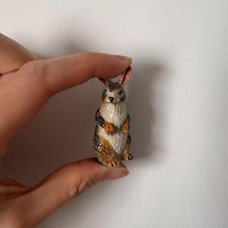 May include: A small, detailed ceramic rabbit figurine. The rabbit has a white chest and belly, with brown and tan markings on its back and sides. It has pink-lined ears and blue eyes. The figurine is held between two fingers.