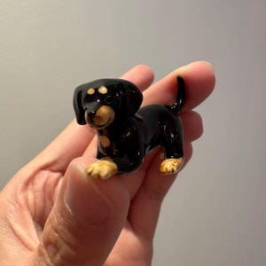 May include: A miniature, glossy black and tan dachshund ornament. The dog has a shiny black coat, tan paws, and a tan face. The ornament is a small representation of a dachshund dog breed.