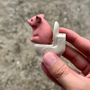 May include: A small, pink pig figurine sitting on a miniature white toilet. The pig is facing left, with its ears perked up. The toilet has a simple design. The figurine is held in a hand.
