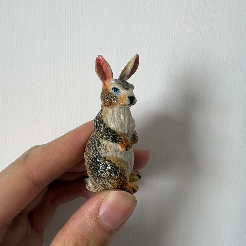 May include: A small, detailed figurine of a rabbit. The rabbit has a mix of brown, tan, and white fur with red-tipped ears and blue eyes. The figurine is held in a hand against a white background.