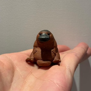 Könnte beinhalten: Eine kleine, braune Schnabeltierfigur mit dunklem Schnabel und rundem Körper. Die Figur sitzt aufrecht, wobei ihre kleinen Füße sichtbar sind. Diese Schnabeltierfigur wäre eine großartige Ergänzung für jede Sammlung.