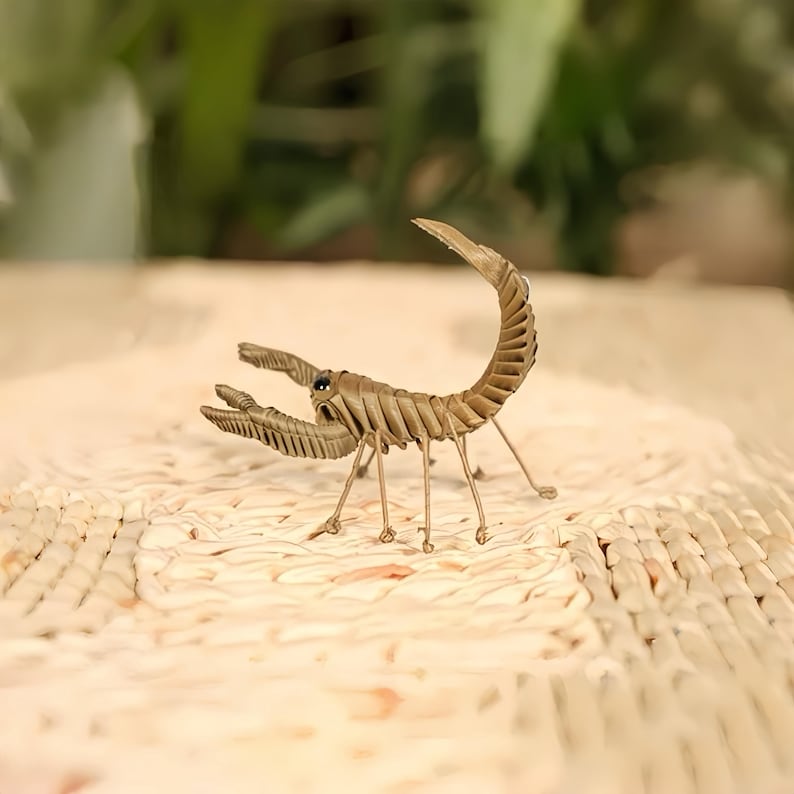 May include: A detailed, handcrafted scorpion sculpture in shades of tan and brown. The scorpion features intricate detailing on its segmented body, claws, and curved tail. The sculpture is posed on a textured surface, with a blurred green background.