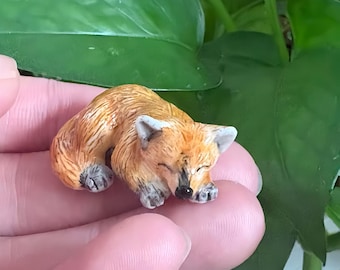 May include: A miniature figurine of a sleeping fox with orange and white fur, pointed ears, and a black nose. The small animal is curled in a resting position with its paws tucked inward, showcasing detailed painted textures on its coat.