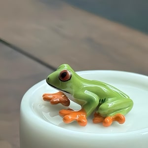 Può includere: Statuetta in ceramica di una rana arboricola. La rana è verde con zampe arancioni e grandi occhi rossi. È posizionata su una superficie bianca e arrotondata, con uno sfondo sfocato.