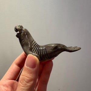 Könnte beinhalten: Eine Keramikfigur eines Seehunds in liegender Position. Der Seehund ist grau mit dunkelgrauen Streifen auf dem Rücken. Die Figur wird in der Hand einer Person gehalten.