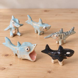 May include: Five small ceramic figurines shaped like sharks with legs. Four are light blue with varying poses and expressions, including one with an open mouth. One figurine is black with white markings, resembling an orca, also with legs.