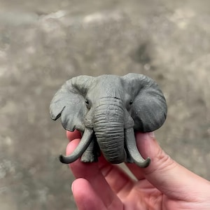 以下が含まれることがあります： 詳細な肌の質感と湾曲した牙を持つ灰色の象の置物。象は大きな耳を持ち、しゃがんだ姿勢をしています。置物は、ぼやけた背景に対して手で持たれています。