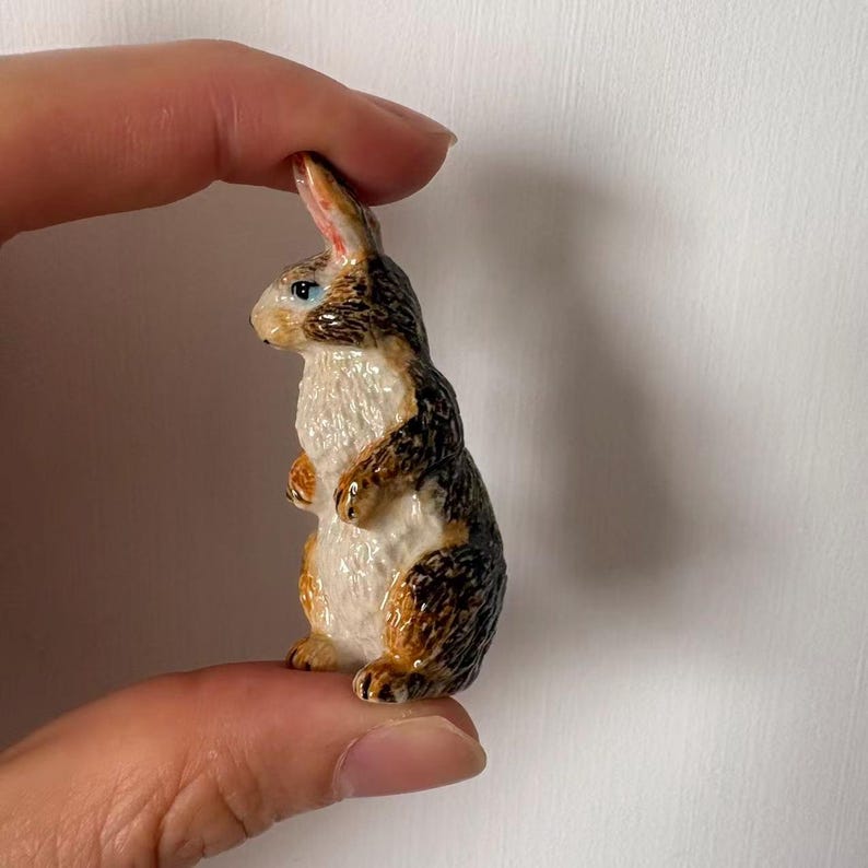 May include: A small ceramic rabbit figurine with a detailed design. The rabbit has a white chest, brown and black markings, and blue eyes. The figurine is held between two fingers.