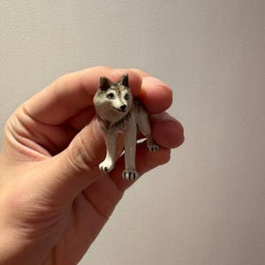May include: A miniature ceramic husky dog figurine. The dog has a white and grey coat, brown ears, and blue eyes. The figurine is held in a hand against a plain background.