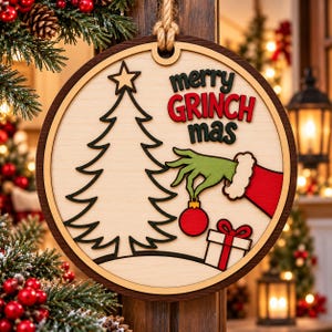 Könnte beinhalten: Ein runder Weihnachtsanhänger aus Holz mit mehrschichtigem Design, bestehend aus einer schwarzen Tannenbaum-Silhouette, einer grünen Hand mit rotem Ärmel, die eine rote Kugel hält, einem weißen Geschenk mit roter Schleife und dem Text merry GRINCH mas.