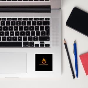 May include: A white laptop computer with a black keyboard and a sticker on the bottom right corner. The sticker is black with a yellow flame and the text "RefinersFire Apparel".