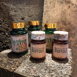 May include: Five vintage pill bottles with gold lids. Two bottles are green with white labels that say "Daytripper" and "Original Buffout". Two bottles are brown with white labels that say "Original Buffout". One bottle is green with a white label that says "Daytrip".