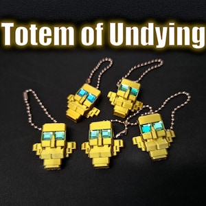 Puede incluir: Cinco llaveros con encanto pixelados de color dorado con detalles azules, cada uno unido a una cadena plateada. Los llaveros están dispuestos sobre una superficie oscura, con el texto "Totem of Undying" arriba.