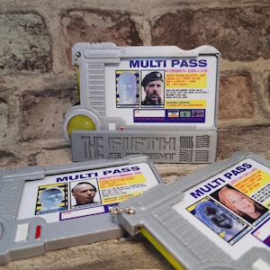 Peut inclure: Trois cartes d'identification rectangulaires en plastique argenté Multi Pass du film Le Cinquième Élément. Chaque carte présente un accent circulaire jaune, une chaînette en métal et une carte d'identité imprimée avec portrait et texte.