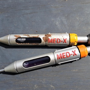Könnte beinhalten: Zwei silberne medizinische Injektoren mit orangefarbenen und gelben Akzenten. Beide Injektoren tragen den Schriftzug "MED-X" in Rot.