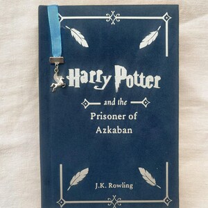 Può includere: Un libro rilegato in blu con un segnalibro argentato raffigurante un cavallo alato. Il titolo del libro è "Harry Potter e il Prigioniero di Azkaban" di J.K. Rowling.