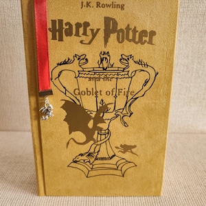 May include: A mustard yellow hardcover book, "Harry Potter and the Goblet of Fire" by J.K. Rowling, with gold embossed title and illustrations. A red ribbon bookmark with a silver charm is attached to the book.