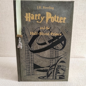 May include: A hardcover book titled "Harry Potter and the Half-Blood Prince" by J.K. Rowling. The book has a dark green cover with gold lettering and a black illustration. A light green ribbon bookmark with a silver charm is attached.