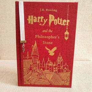Könnte beinhalten: Ein rotes Hardcover-Buch mit dem Titel "Harry Potter and the Philosopher's Stone" von J.K. Rowling. Das Cover zeigt goldene Illustrationen von Hogwarts und einem Vogel. Ein weißes Lesezeichen mit Blitz-Anhänger ist eingelegt.