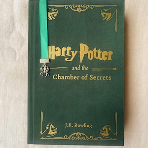 Può includere: Un libro verde rilegato con un segnalibro dorato e un ciondolo a forma di ragno argentato. Il titolo del libro è "Harry Potter e la Camera dei Segreti" di J.K. Rowling.