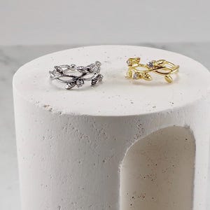 May include: Two rings, one silver and one gold, each with a detailed leaf and vine pattern. The silver ring has clear gemstone details, and the gold ring has similar accents. Both rings are presented on a white, textured surface.