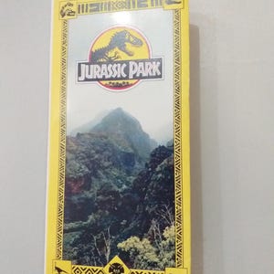 Könnte beinhalten: Eine VHS-Kassette für den Film "Jurassic Park". Das Cover zeigt das Logo des Films mit einer T-Rex-Silhouette in einem Kreis, vor einer Berglandschaft. Der Rand ist gelb mit dekorativen Mustern.