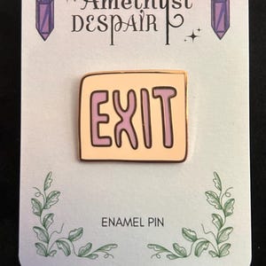 May include: An enamel pin on a white card with the text "EXIT" in a light orange color with a brown outline. The card has the text "Amethyst Despair" and "ENAMEL PIN" with purple crystal and leaf illustrations.