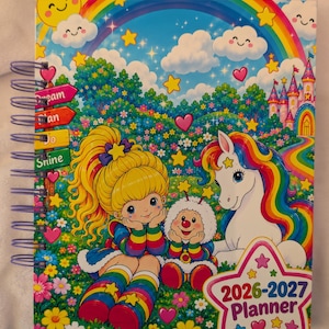 May include: A spiral-bound 2026-2027 planner featuring a colorful illustration of a character with blonde hair, a small creature, and a unicorn in a flower field under a rainbow. The cover includes signs labeled Dream, Plan, Do, and Shine.