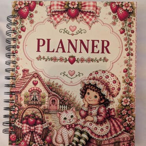 May include: A spiral-bound planner featuring a cream-colored cover with a central frame containing the word PLANNER. The design includes a cottage, a child in a strawberry-patterned outfit, a cat, a basket of strawberries, and floral borders.