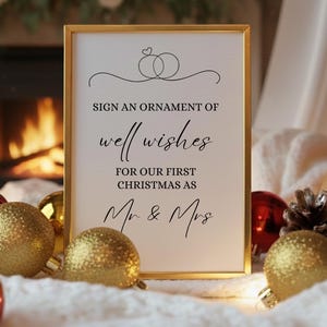 May include: A framed sign with gold trim displays the text "Sign an ornament of well wishes for our first Christmas as Mr & Mrs." The sign is surrounded by gold and red Christmas ornaments and a white blanket.