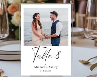 May include: Clear acrylic table number with a photo of a couple holding hands. The text "Table 8" is written in a script font, with the names "Michael + Ashley" and the date "3. 1. 2026" below.