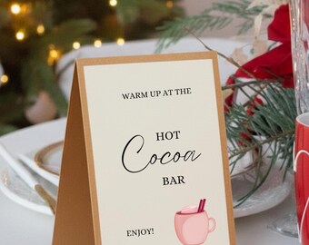 May include: A folded sign with a light beige background and a brown border. The sign reads "Warm up at the Hot Cocoa Bar. Enjoy!" A pink mug of hot cocoa with whipped cream and cinnamon sticks is illustrated on the sign. The sign is on a white surface.