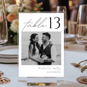 Può includere: Numero tavolo 13 in acrilico trasparente. Presenta una foto in bianco e nero di una coppia, con la scritta "table 13" in alto. I nomi "Michael & Ashley" e la data "03 OTTOBRE 2027" sono in basso.